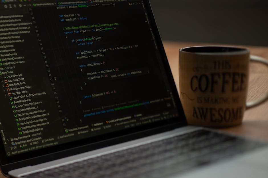Close-up view of a laptop screen displaying lines of code in a dark-themed text editor, with colorful syntax highlighting. To the right of the laptop, there is a ceramic coffee mug with black text tha